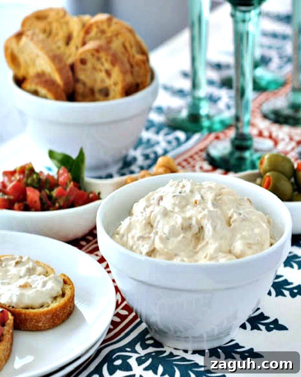 a white serving bowl of caramelized shallot dip, with toasted baguette slices, a bowl of olives, and green stemmed wine glasses in background