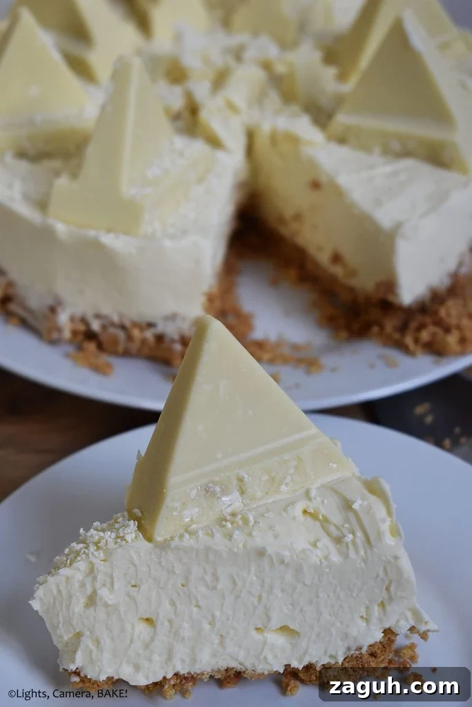 White Chocolate Toblerone Cheesecake Recipe — Creamy No-Bake Slice 7 Vertical view of a slice of White Chocolate Toblerone Cheesecake on a white plate with the full cheesecake in the background on a brown board.