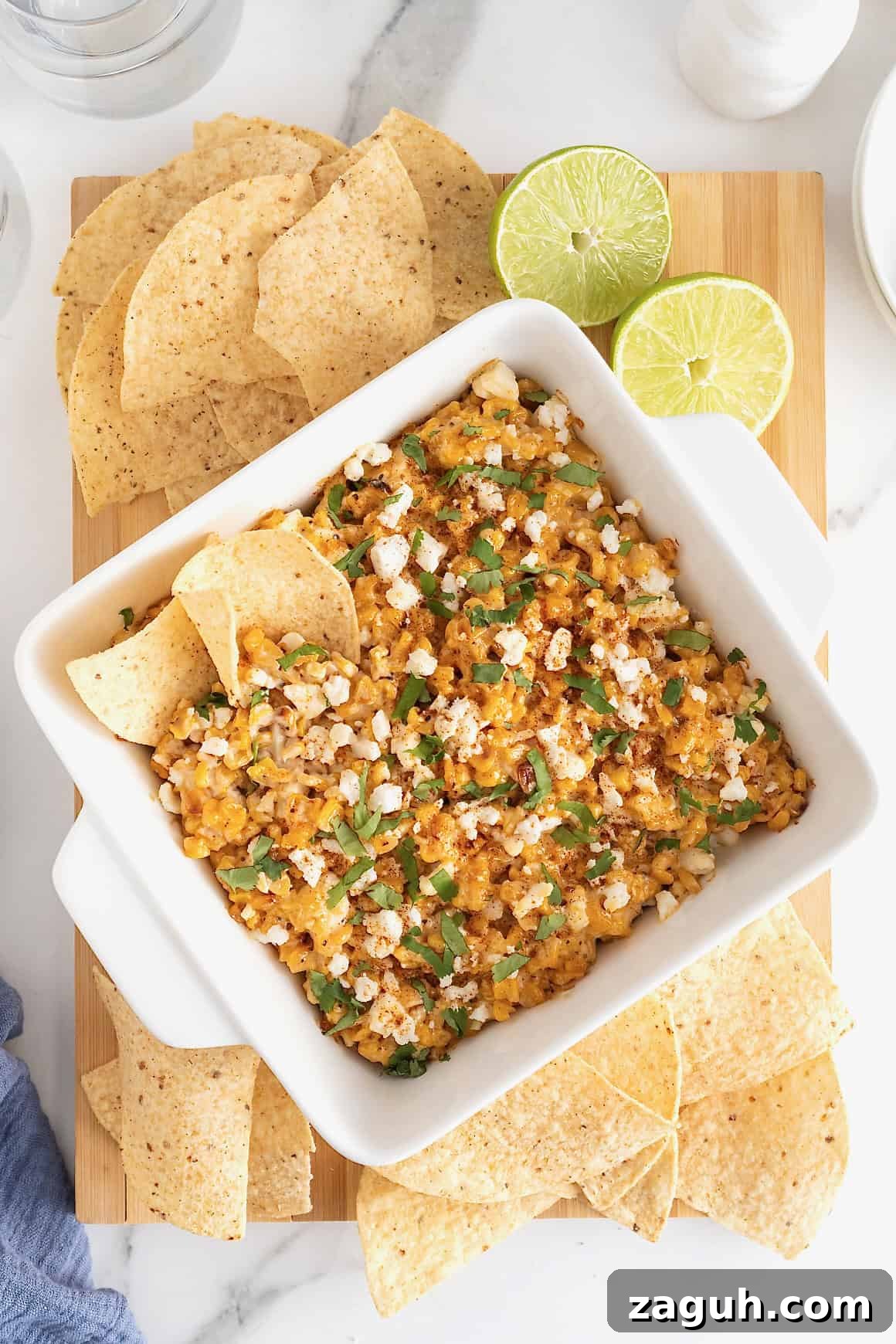 Authentic Mexican Fiesta Recipes for Your Party Menu 5 A square white casserole dish of elotes dip surrounded by corn chips on a wooden cutting board.