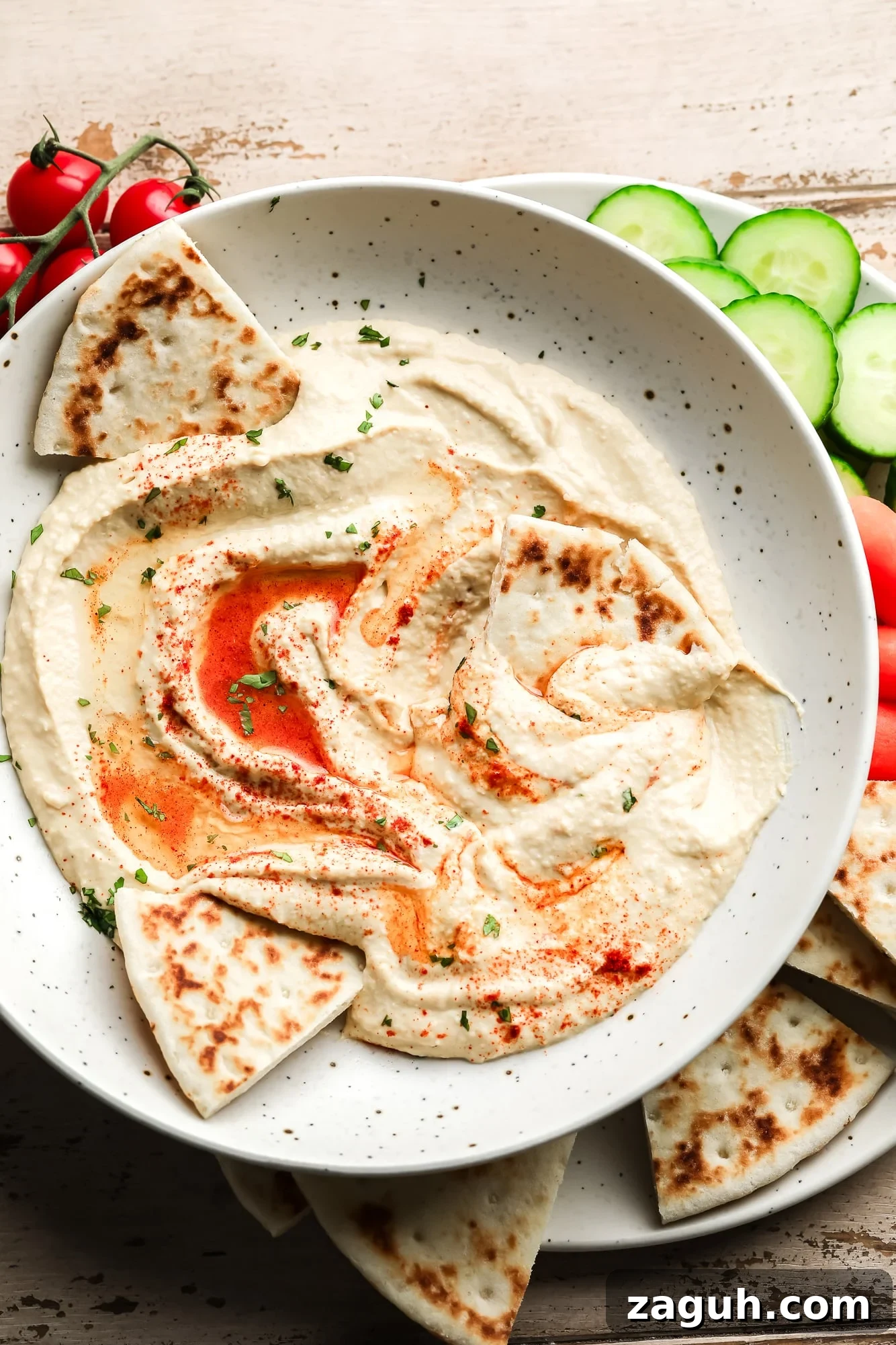 hummus topped with paprika and oil in a white bowl with pita bread and vegetables on the side.