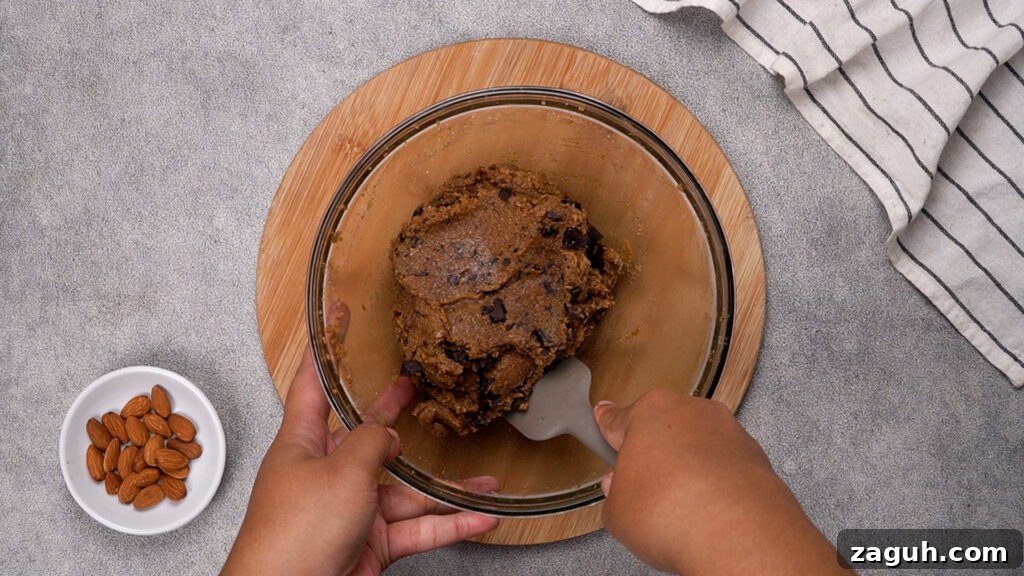 Almond Flour Chocolate Chip Cookie Recipe — Soft & Chewy</assistant 8 Hands folding and mixing the cookie dough with a spatula, making sure the chocolate is evenly distributed.