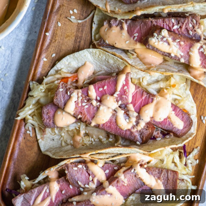 Overhead view of four seared tuna tacos on a wooden serving board garnished with sriracha mayo and sesame seeds.