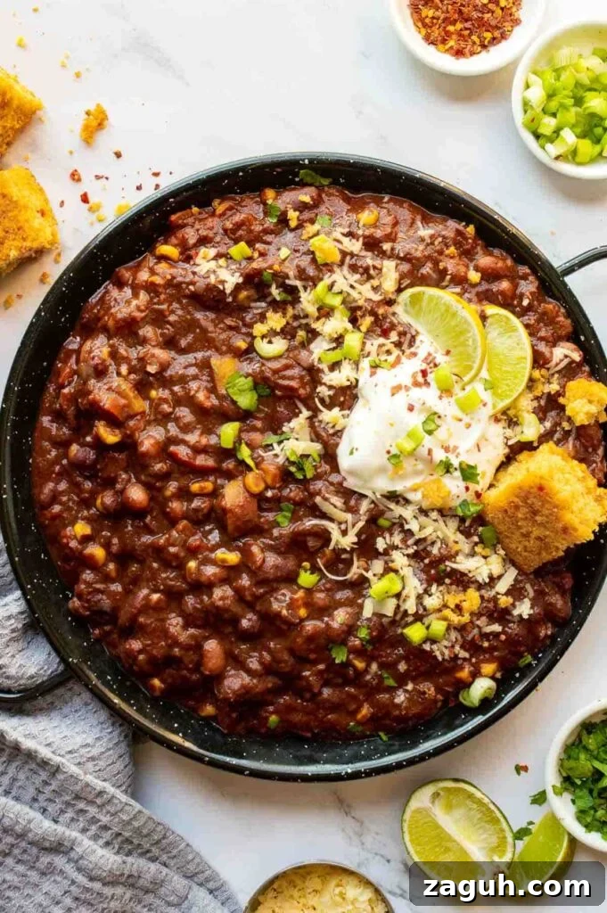 Guinness Vegan Chili Recipe: Rich, Hearty Stovetop Chili 2 Top-down view of a large black pan with chili topped with cornbread and white sauce and lime slices, on a white background surrounded by chopped herbs and ground spices.