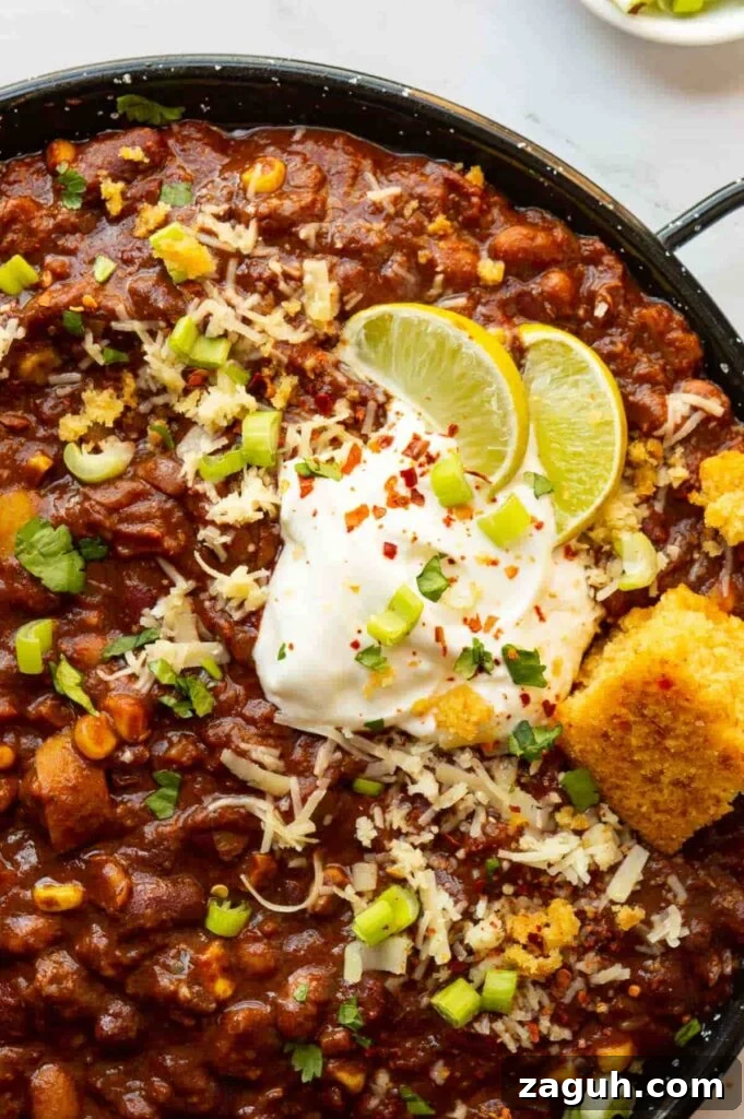 Guinness Vegan Chili Recipe: Rich, Hearty Stovetop Chili 4 Top-down close-up of a black pan with bean chili topped with sliced limes, white sauce and cornbread.