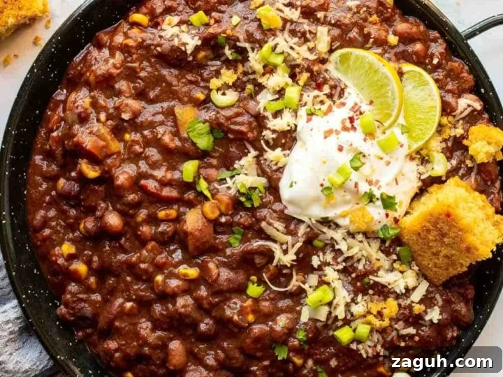 Top-down view of a large black pan with chili topped with cornbread and white sauce and lime slices, on a white background surrounded by chopped herbs and ground spices.