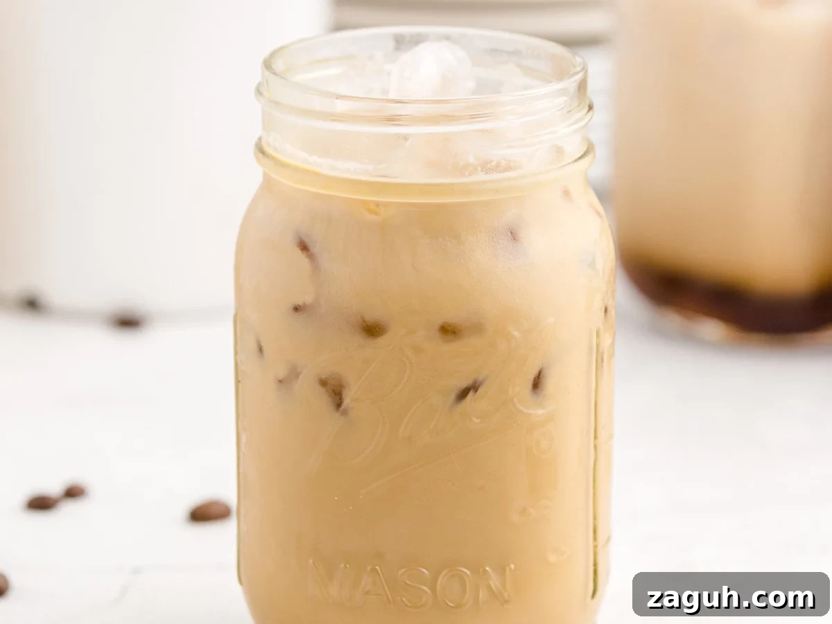 A mason jar filled with Iced Caramel Macchiato sits on a white surface. Ice cubes glisten in the light brown drink, coffee beans are scattered nearby, and another glass of this Iced Caramel Macchiato recipe is blurred in the background.