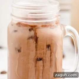 A mason jar mug filled with iced chocolate milk, featuring chocolate syrup swirls and ice cubes, evokes the look of an Iced Caramel Macchiato recipe against a light blurred background.