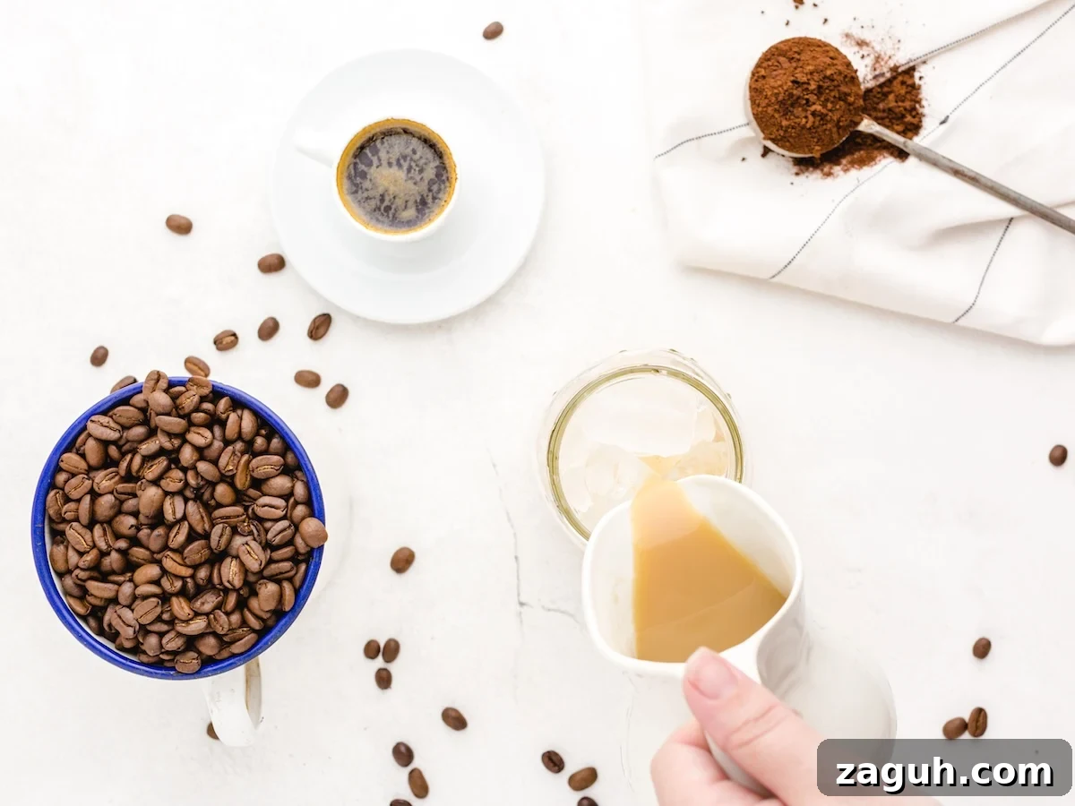 A hand pours milk into a glass with ice, surrounded by a cup of coffee beans, black coffee, a spoon with ground coffee, and scattered beans—perfect inspiration for your next Iced Caramel Macchiato or homemade Iced Caramel Macchiato recipe.