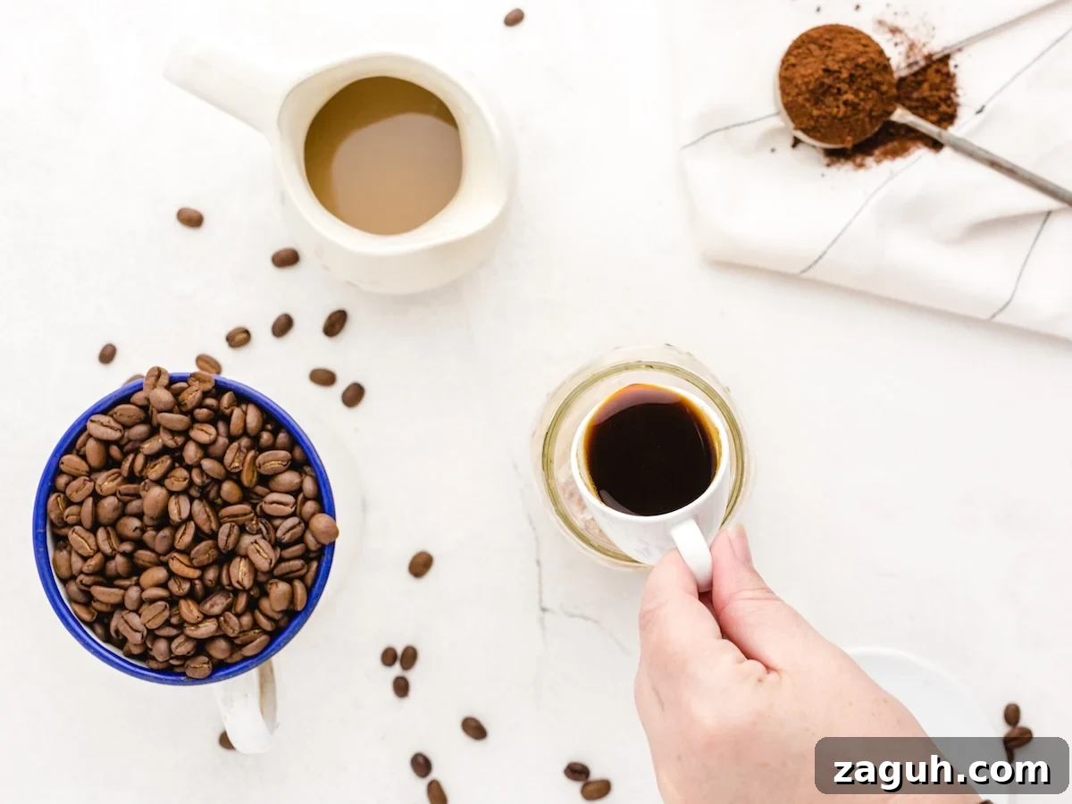 A hand holds a cup of black coffee on a white surface. Nearby are a mug of beans, creamer, scattered beans, and a spoon with grounds—perfect inspiration for your next Iced Caramel Macchiato Recipe.