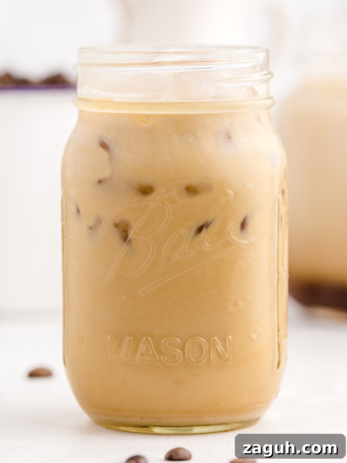 A mason jar labeled Ball Mason is filled with a refreshing Iced Caramel Macchiato and ice cubes, sitting on a light surface with coffee beans scattered nearby—perfect inspiration for your next Iced Caramel Macchiato recipe.