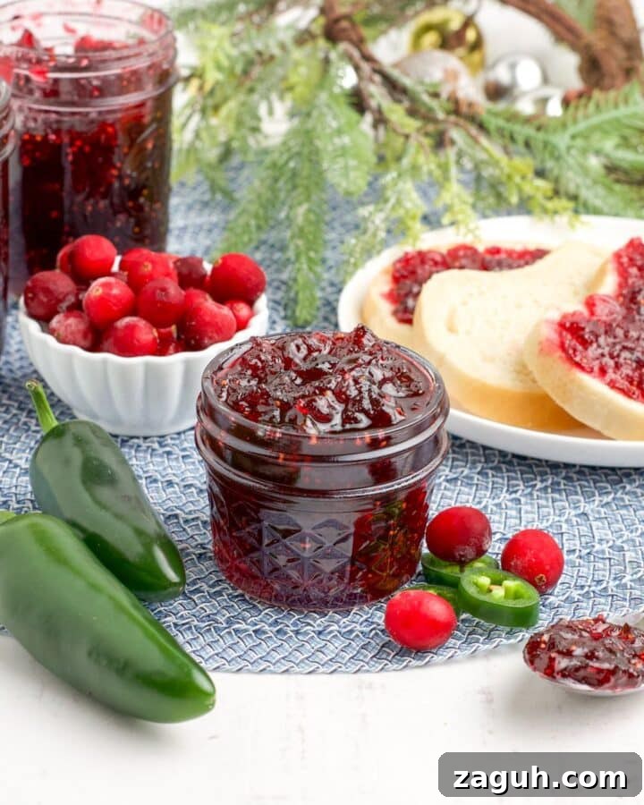 A jar of cranberry jalapeno jam on the table with a spoon to the side and a bowl of cranberries in the background.