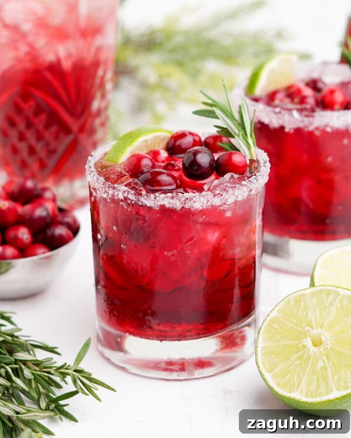 Cranberry margarita on a white table with fresh lime, rosemary and cranberries.