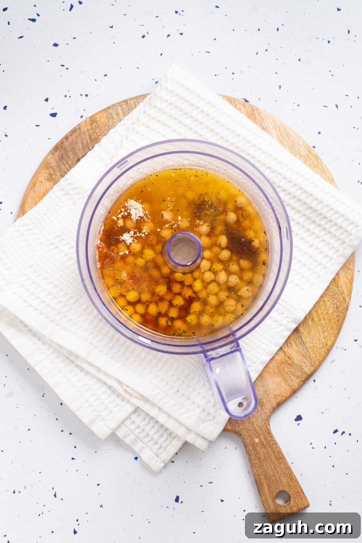 Overhead view of chickpeas, oil, nutritional yeast, soy sauce, onion powder, garlic powder, poultry seasoning, and miso paste inside the food processor. 