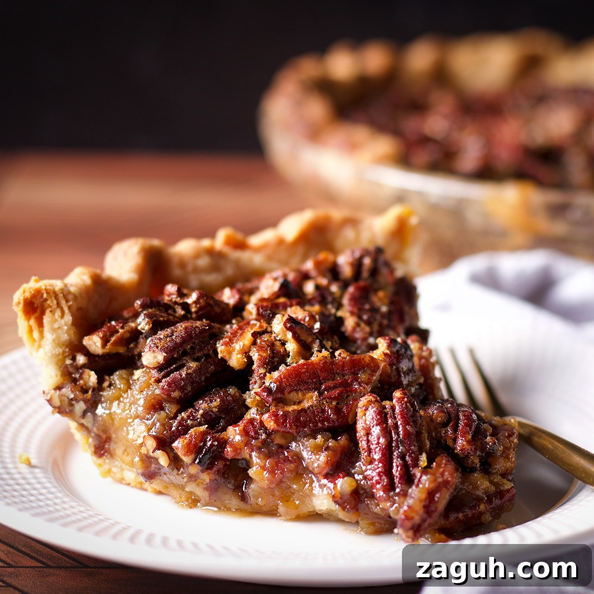 Pecan Pie Made Without Corn Syrup — Classic Recipe with Pure Maple 5 A slice of pecan pie on a white plate with a gold fork resting next to it.