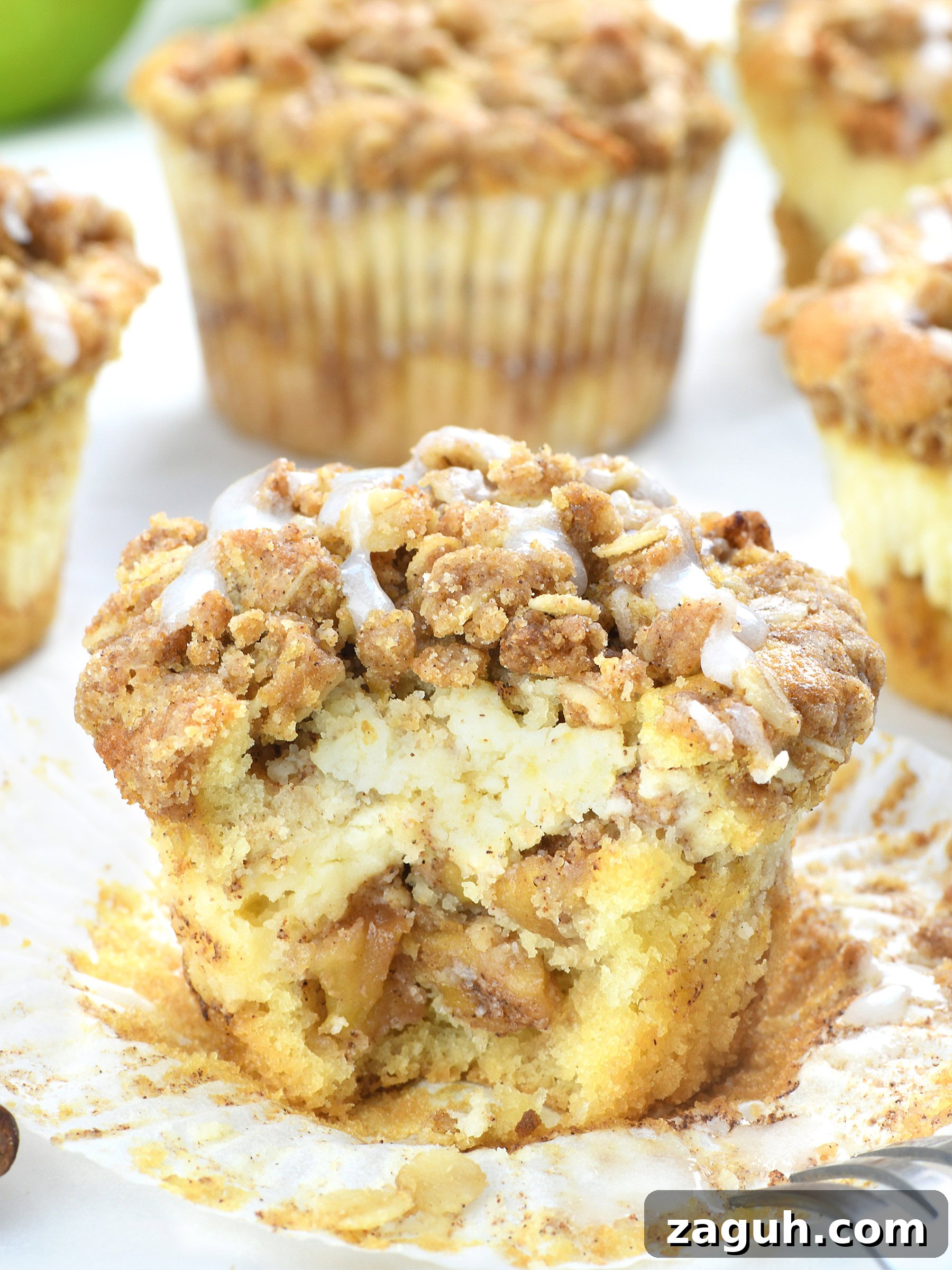 Apple Crisp Cheesecake Muffins with Cinnamon Streusel 2 Close-up of an apple crisp cheesecake muffin with a crumbly cinnamon-oat topping, creamy cheesecake center, and tender apple filling, drizzled with vanilla glaze.