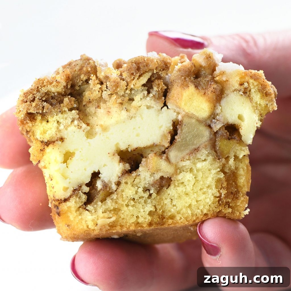 Apple Crisp Cheesecake Muffins with Cinnamon Streusel 9 Hand holding half of an apple crisp cheesecake muffin showing layers of moist cake, creamy cheesecake filling, spiced apple chunks, and a crumbly cinnamon streusel topping.
