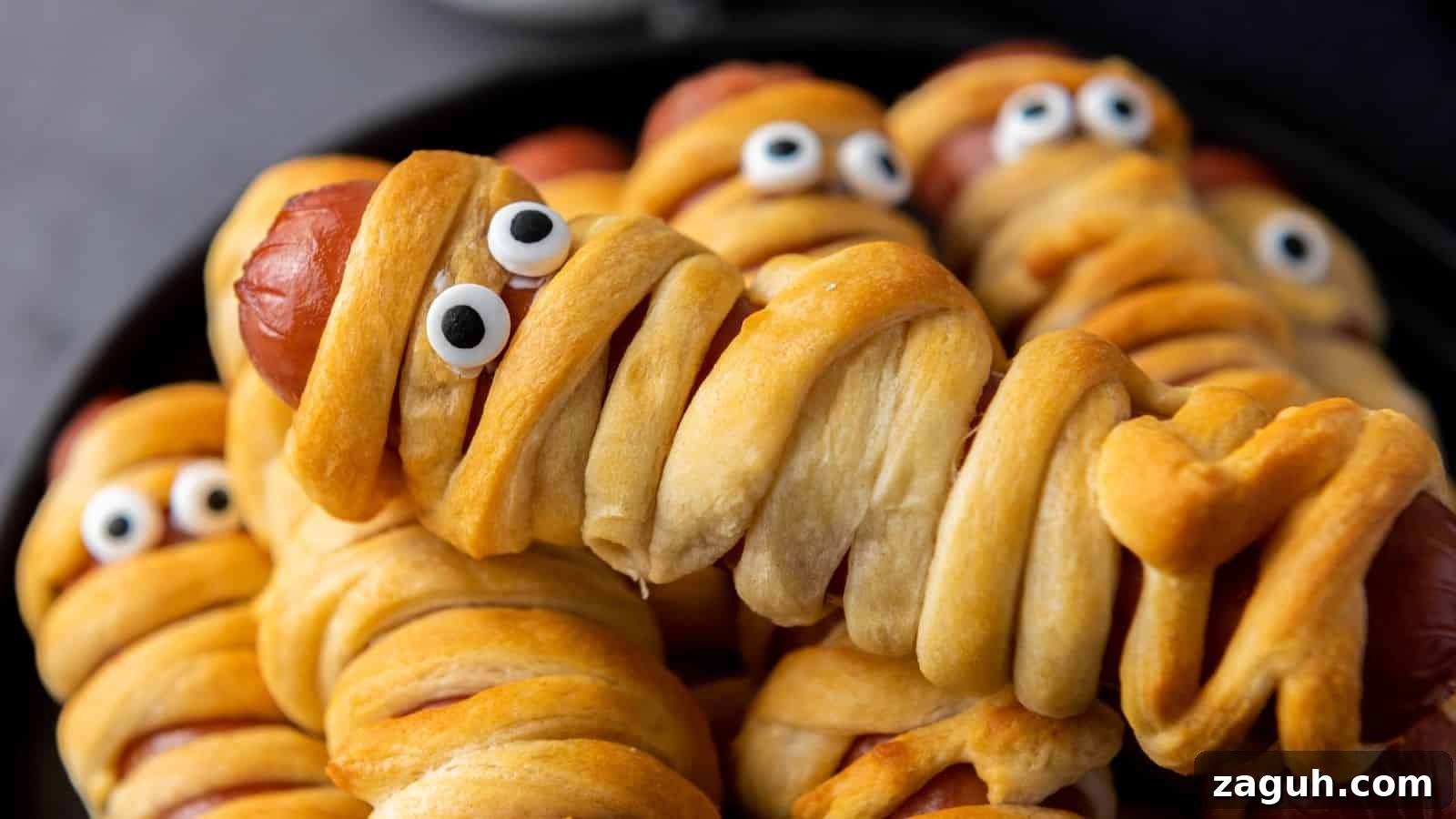 Hot dogs wrapped in dough to resemble mummies, complete with candy eyes, arranged on a black plate.