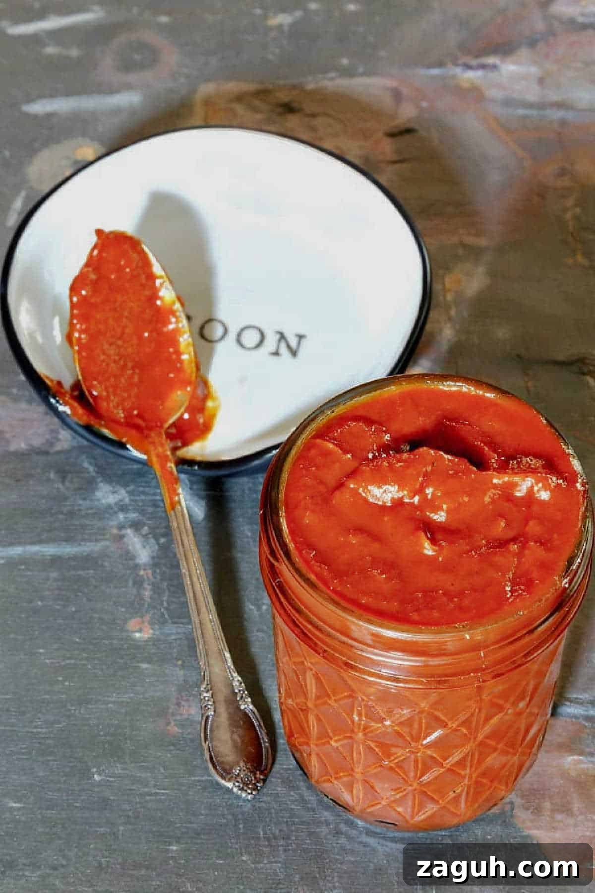 A glass canning jar of spicy ketchup with some ketchup on a spoon on a spoon rest.