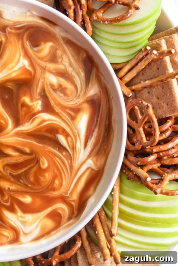 Caramel cream cheese dip swirled in a white bowl next to pretzels, graham crackers, and green apples.
