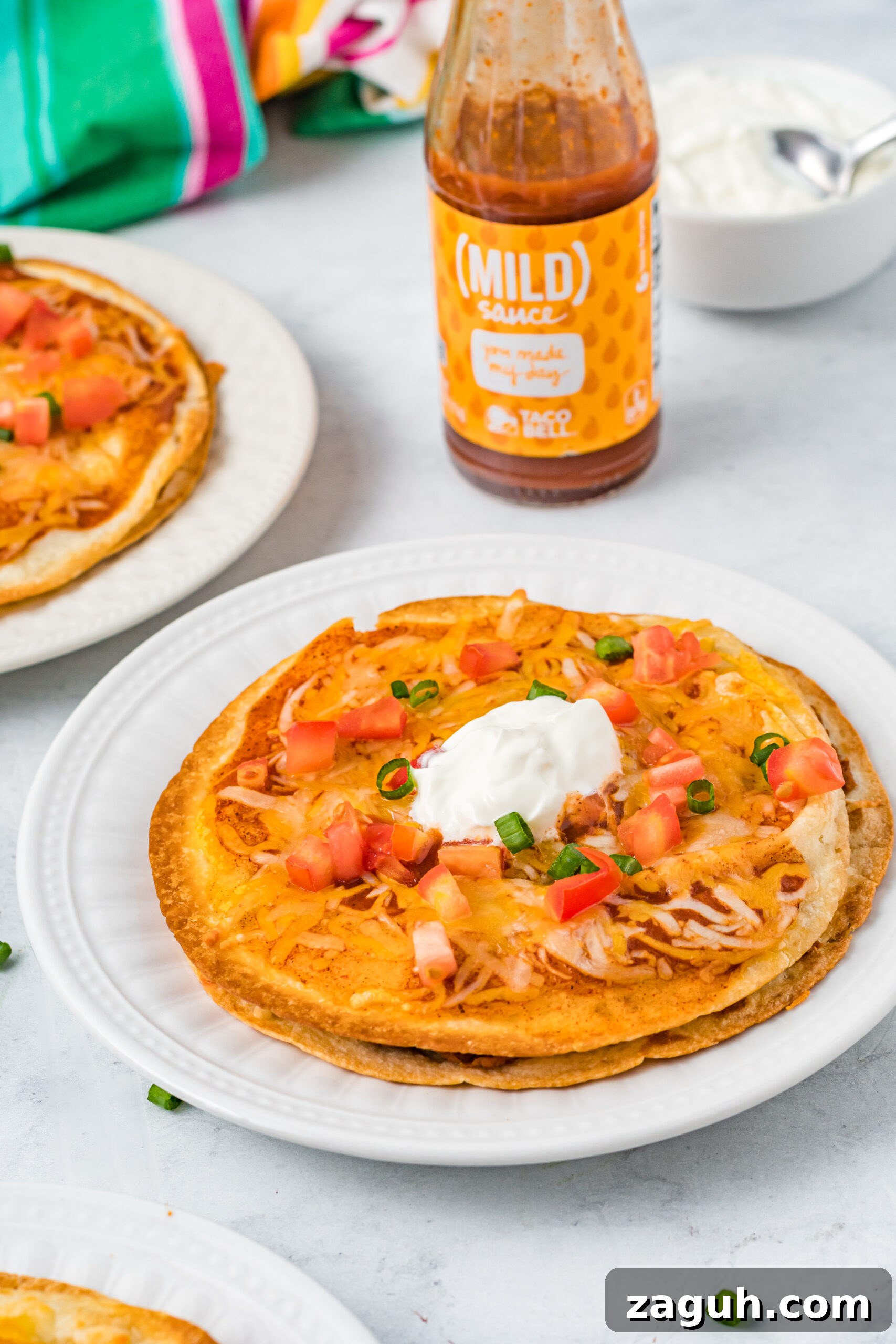 Taco Bell Mexican Pizza: How to Order, Ingredients & Nutrition Facts 13 Mexican Pizza on a white plate topped with sour cream, tomatoes, and green onion.