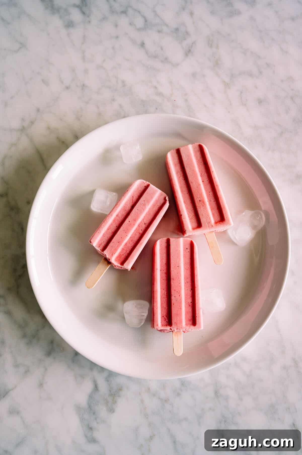 Strawberry Greek Yogurt Popsicles Recipe — Creamy Frozen Treats 2 three homemade strawberry popsicles on a white plate with a few ice cubes.