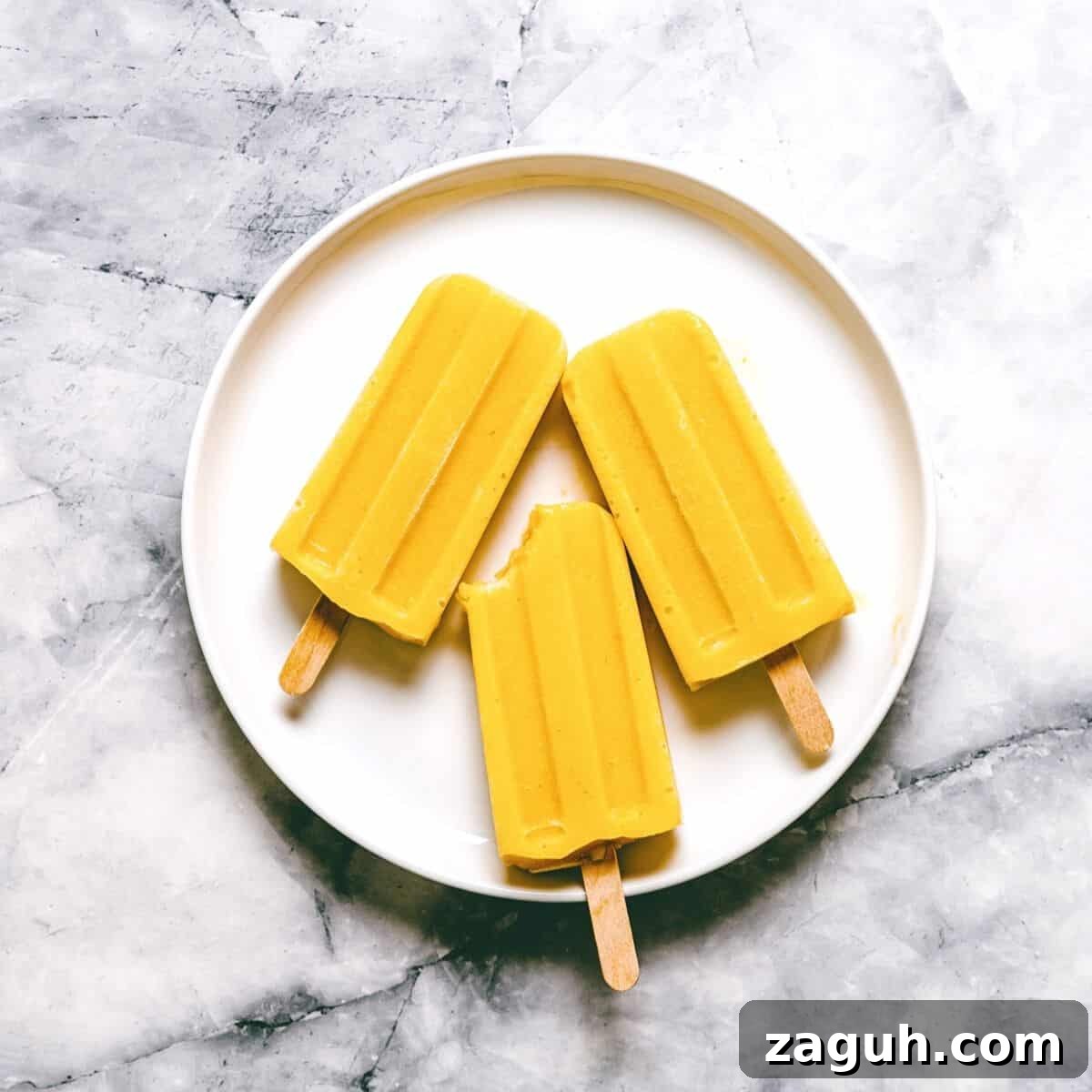 3 mango popsicles on a white plate on a marble surface.