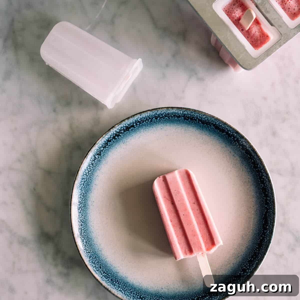 Strawberry Greek Yogurt Popsicles Recipe — Creamy Frozen Treats 9 a strawberry popsicle on a white plate with a blue rim.