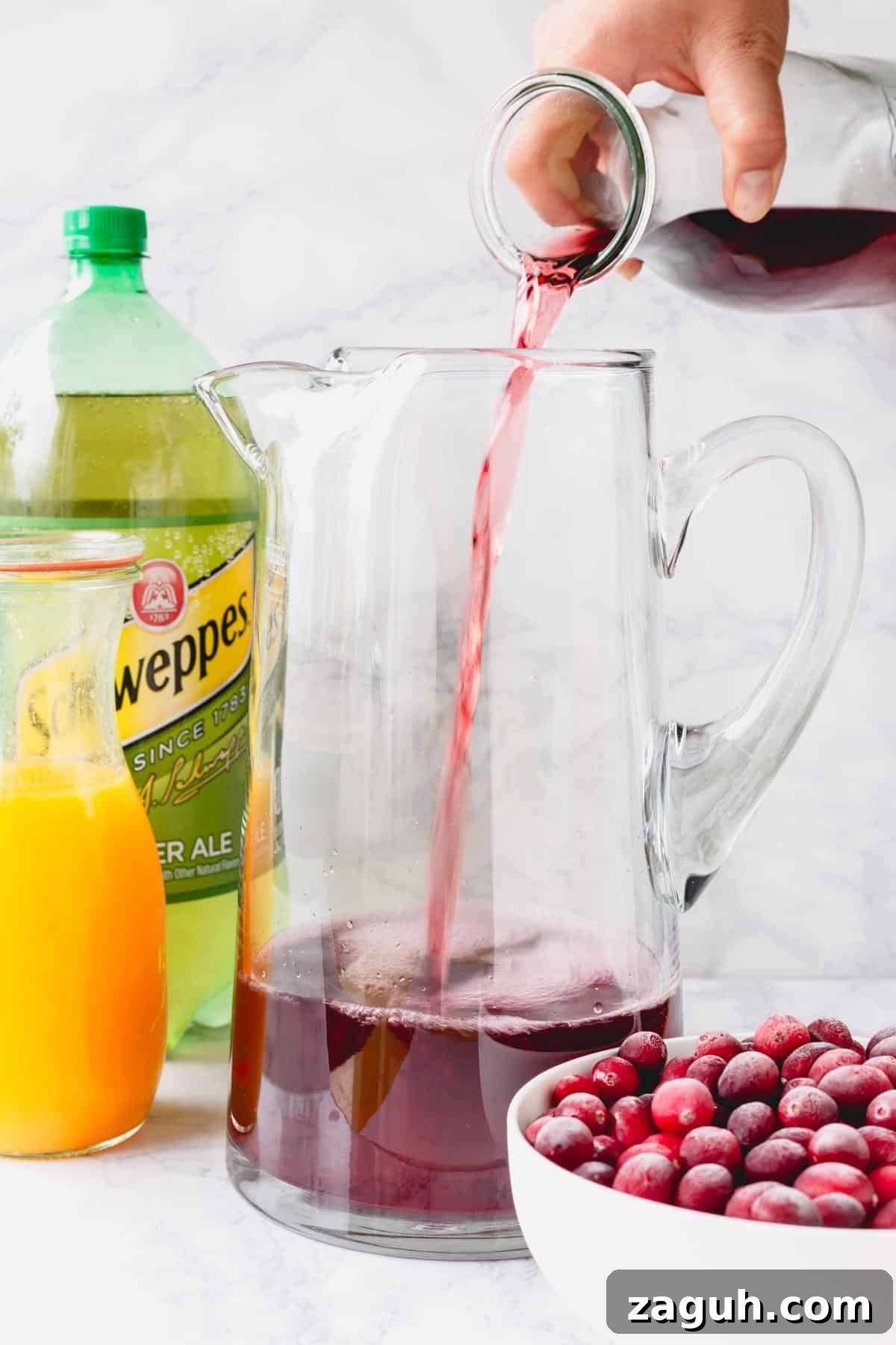 Sparkling Orange Cranberry Spritzer Recipe for Holiday Parties 5 Cranberry juice being poured into a pitcher with ginger ale, orange juice, and pomegranates next to it.