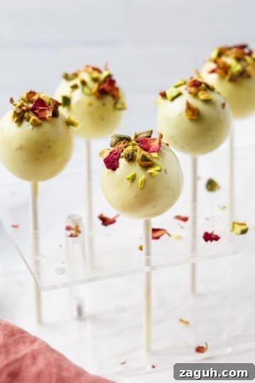 Pistachio cake pops coated in white chocolate topped with chopped pistachios and dried rose petals displayed in acrylic cake pop stand