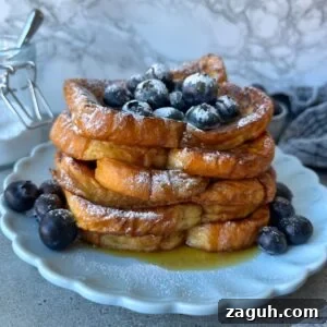 Decadent Brioche French Toast Recipe for Brunch and Dessert 9 A stack of Brioche French Toast slices topped with blueberries and powdered sugar on a white plate, with syrup and extra blueberries around the base.