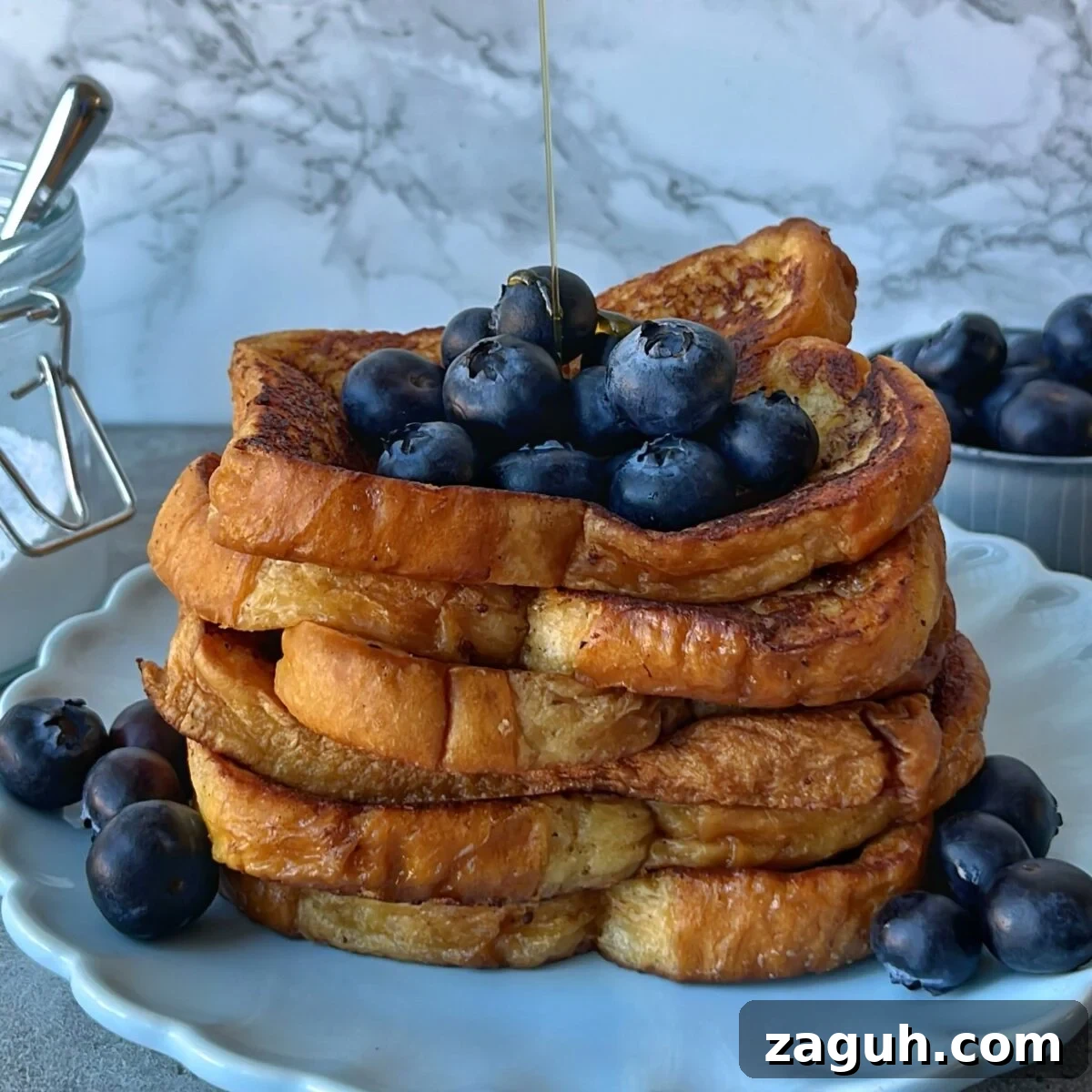 Decadent Brioche French Toast Recipe for Brunch and Dessert 3 A stack of Brioche French Toast topped with fresh blueberries, with syrup being poured over, served on a white plate.