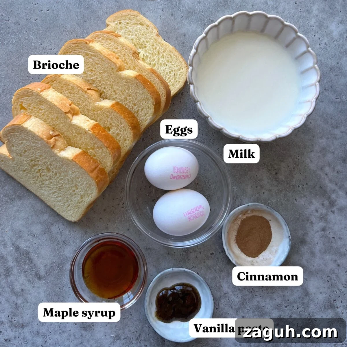 Decadent Brioche French Toast Recipe for Brunch and Dessert 4 Brioche French Toast ingredients: Sliced brioche, two eggs, a bowl of milk, cinnamon, maple syrup, and vanilla paste arranged on a gray surface with labels.