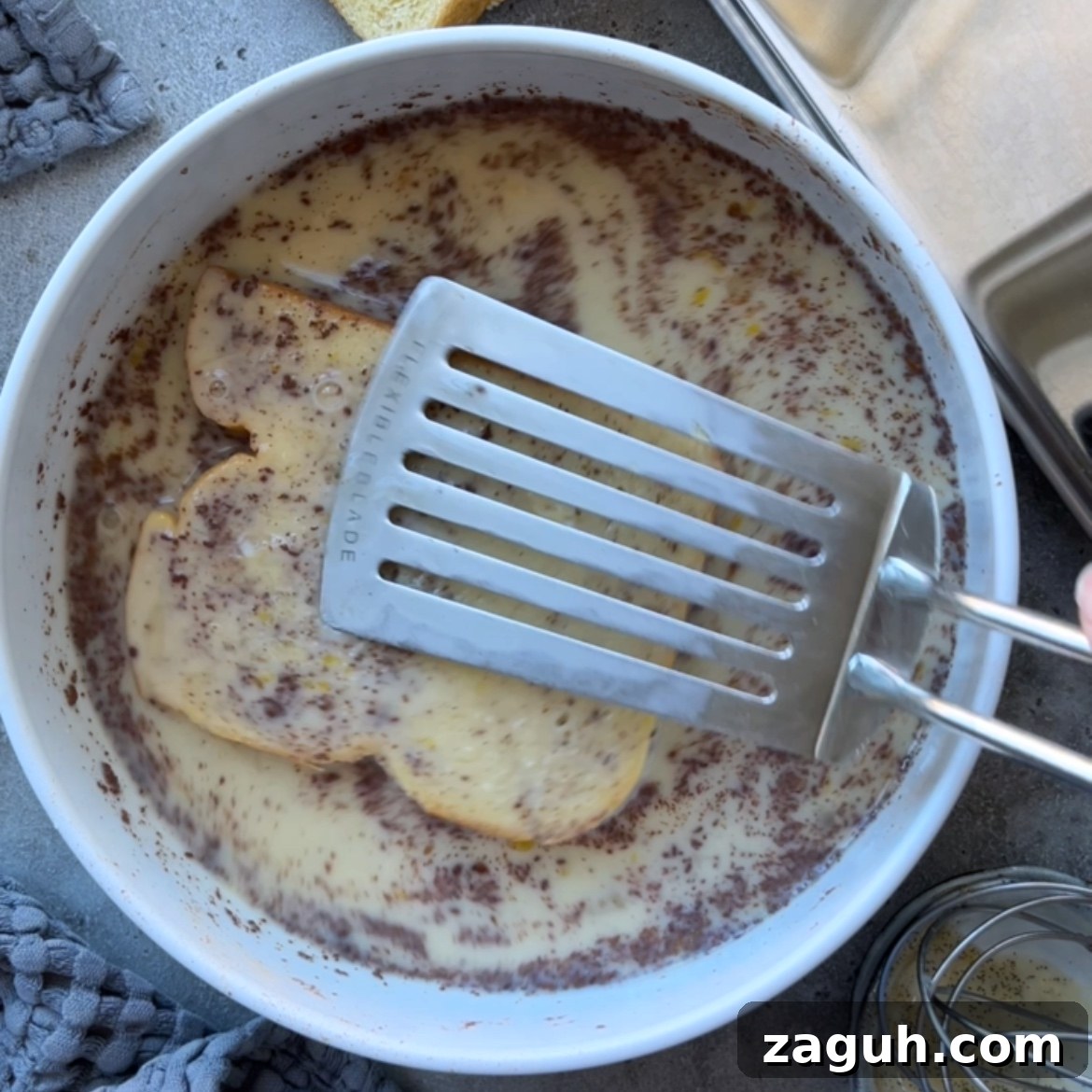 Decadent Brioche French Toast Recipe for Brunch and Dessert 6 A metal spatula pushes sliced brioche into a white dish filled with an egg and milk mixture.