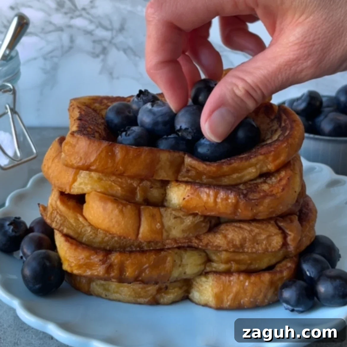 Decadent Brioche French Toast Recipe for Brunch and Dessert 8 A hand places fresh blueberries atop a stack of Brioche French Toast slices on a light blue plate, with more blueberries scattered around.