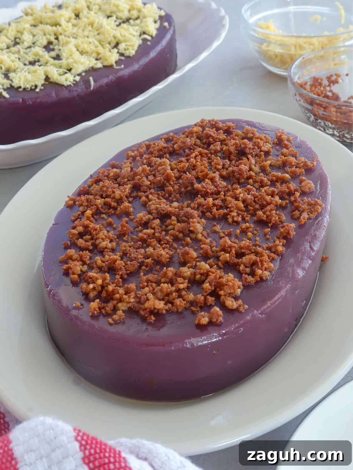 Ube Halaya Recipe: How to Make Creamy Filipino Purple Yam Dessert 2 ube halaya topped with latik on a white serving platter with a ube halaya topped with cheese in the background.