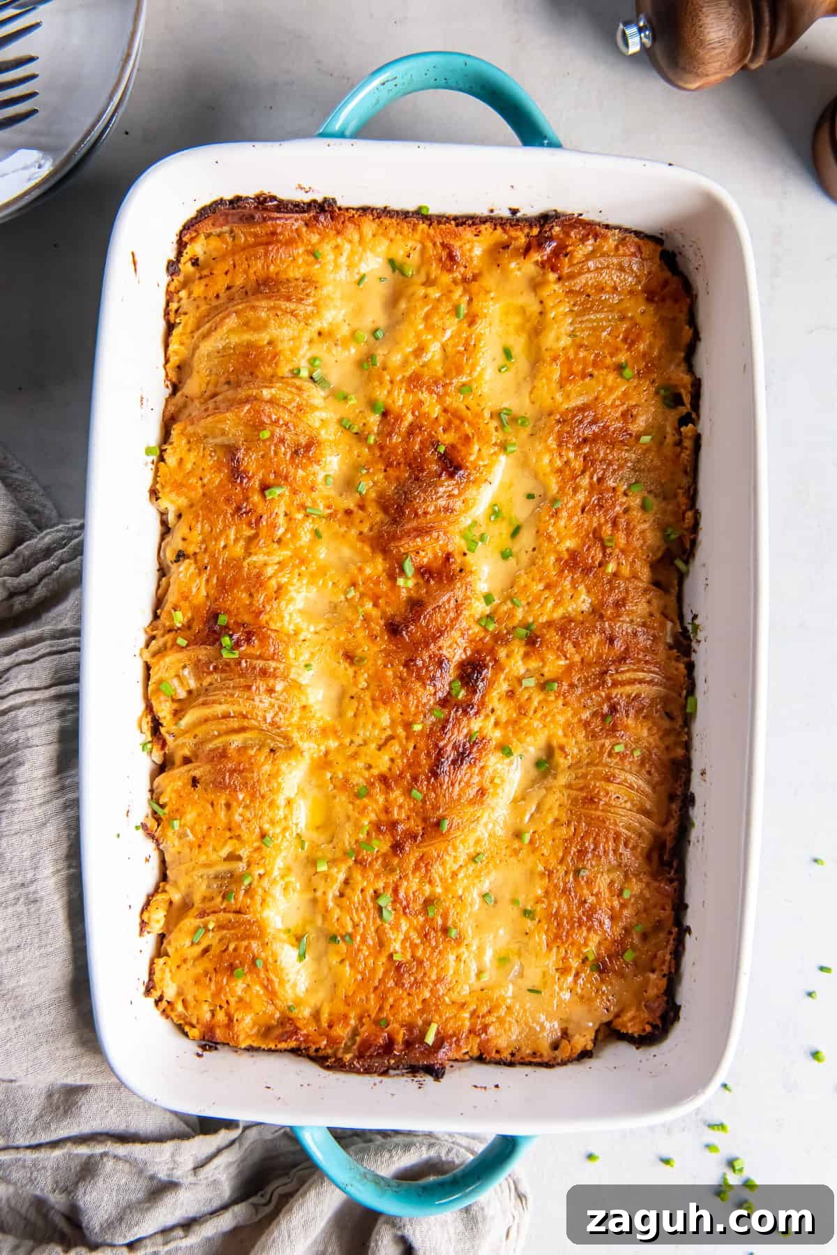 Creamy Scalloped Potatoes au Gratin Recipe 2 A top down shot of au gratin potatoes in a baking dish with blue handles next to a grey cloth.