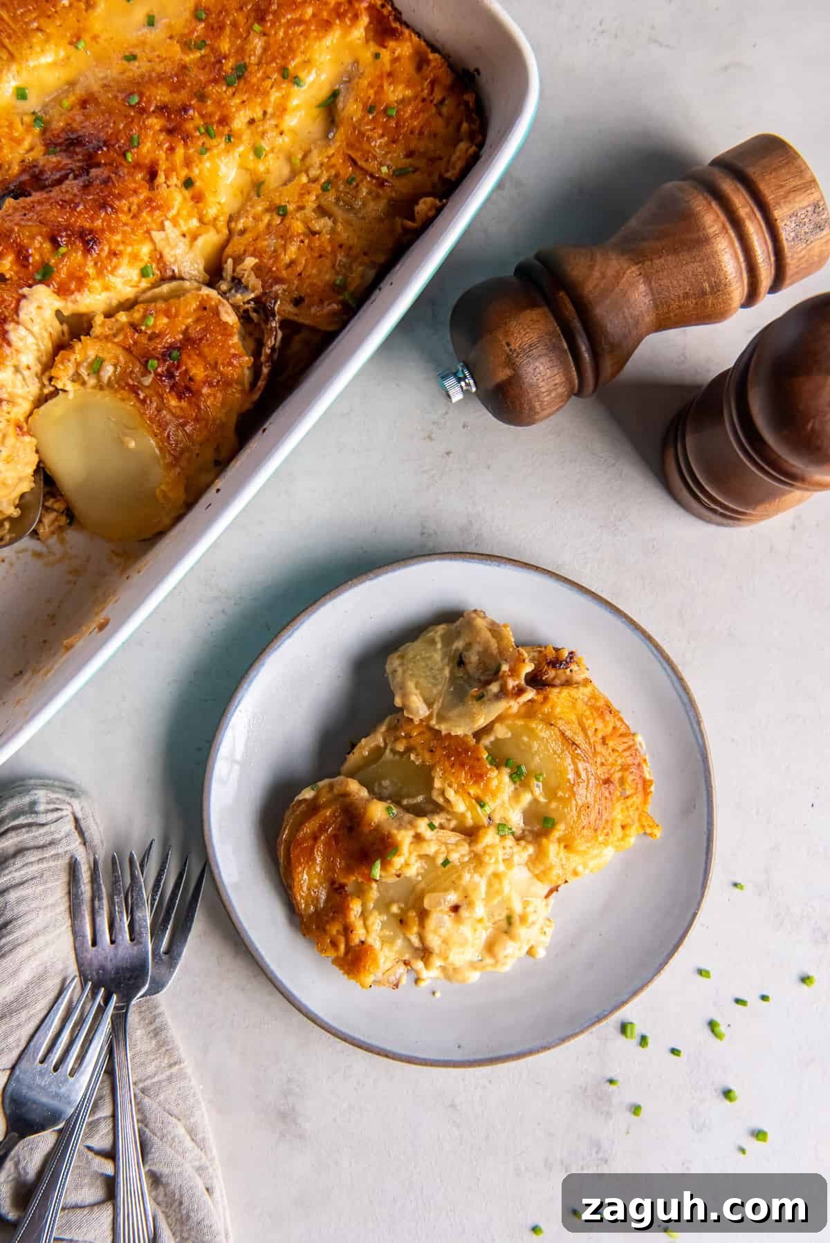 Creamy Scalloped Potatoes au Gratin Recipe 10 A top down shot of a serving of cheesy au gratin potatoes on a white plate next to salt and pepper shakers and a baking dish full of the potatoes.