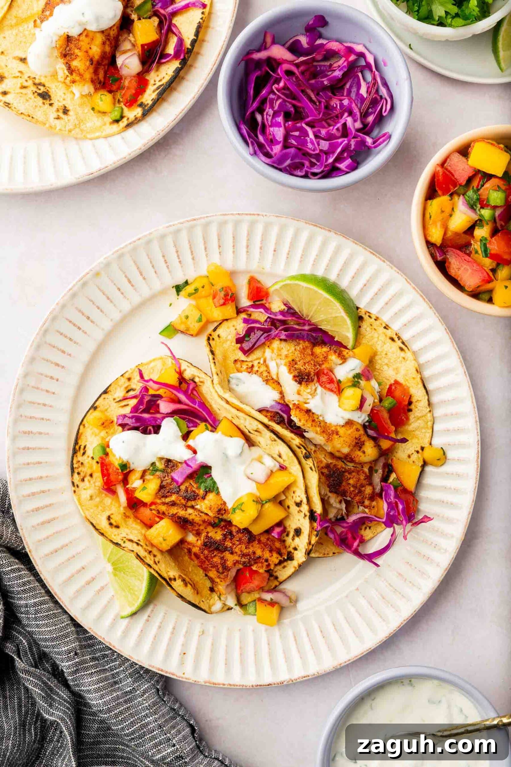 Crispy Tilapia Fish Tacos with Zesty Lime Slaw 4 Two fish tacos on charred corn tortillas topped with sour cream, fruit salsa, and red cabbage with bowls of slaw, fruit salsa, and crema to the side.
