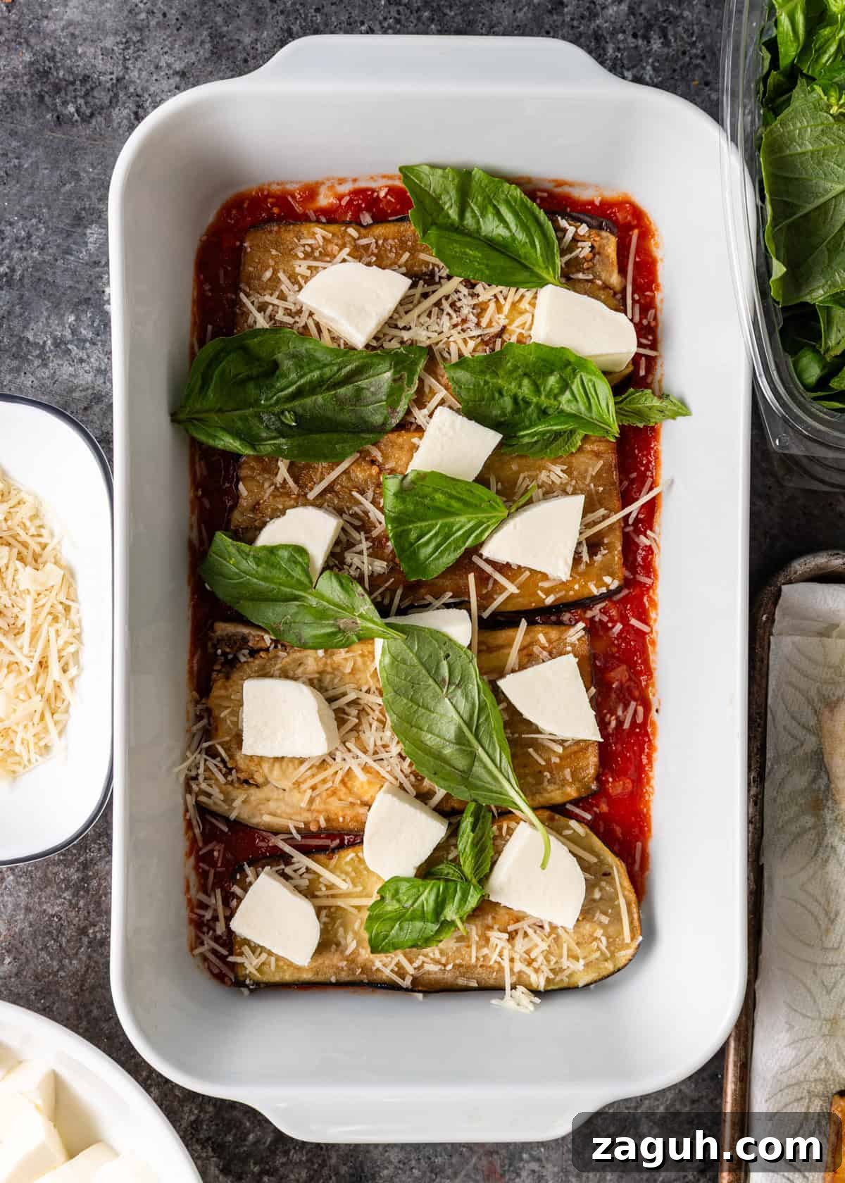 Classic Eggplant Parmigiana Recipe: Authentic Italian Parmigiana di Melanzane 11 overhead process shot: white baking dish with sauce on bottom, layered with egg plant and cut mozzarella, basil and parmesan cheese on top