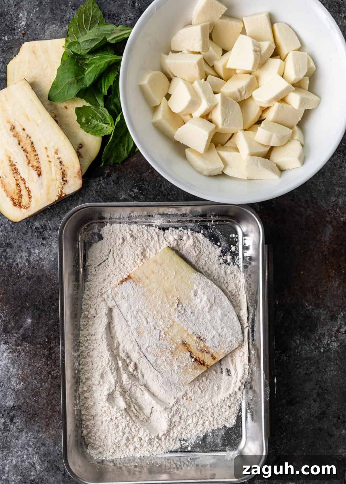 Classic Eggplant Parmigiana Recipe: Authentic Italian Parmigiana di Melanzane 7 overhead process shot: two slices of eggplant on counter with basil besides bowl of cut mozzarella and pan with flour covering a slice of eggplant
