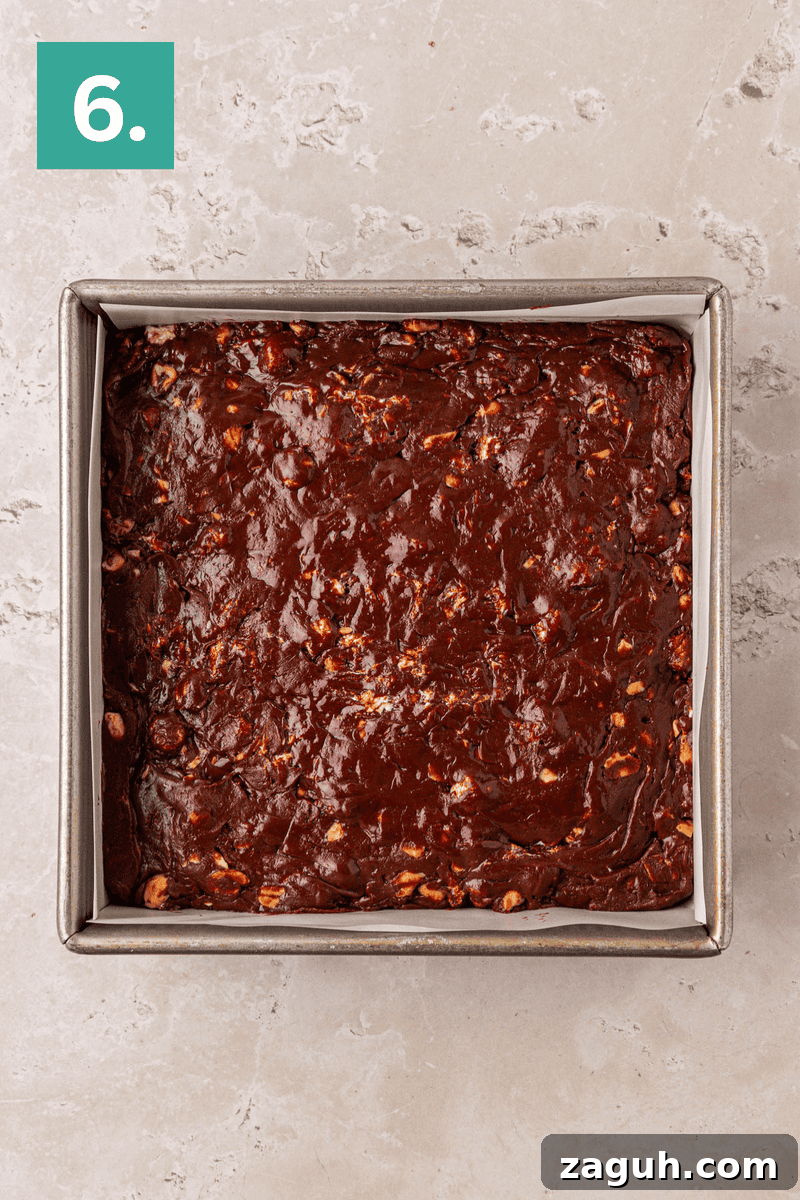 A square metal baking pan filled with unbaked chocolate brownie batter, resting on a light-colored, textured countertop. A green square with the number 6 is in the top left corner.