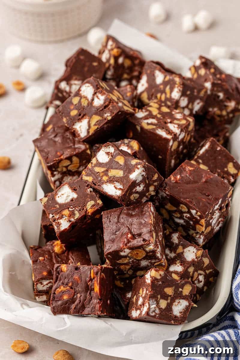 A tray filled with squares of rocky road fudge, showing chunks of marshmallows and peanuts inside the rich chocolate. Some peanuts and mini marshmallows are scattered around the tray.