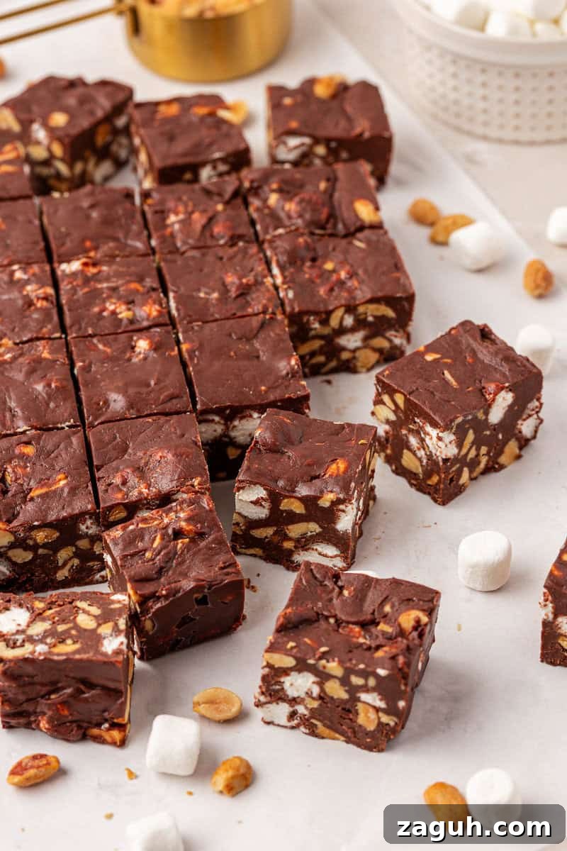 Squares of chocolate fudge with visible peanuts and mini marshmallows are arranged on parchment paper, with some pieces separated. Loose peanuts and mini marshmallows are scattered around.