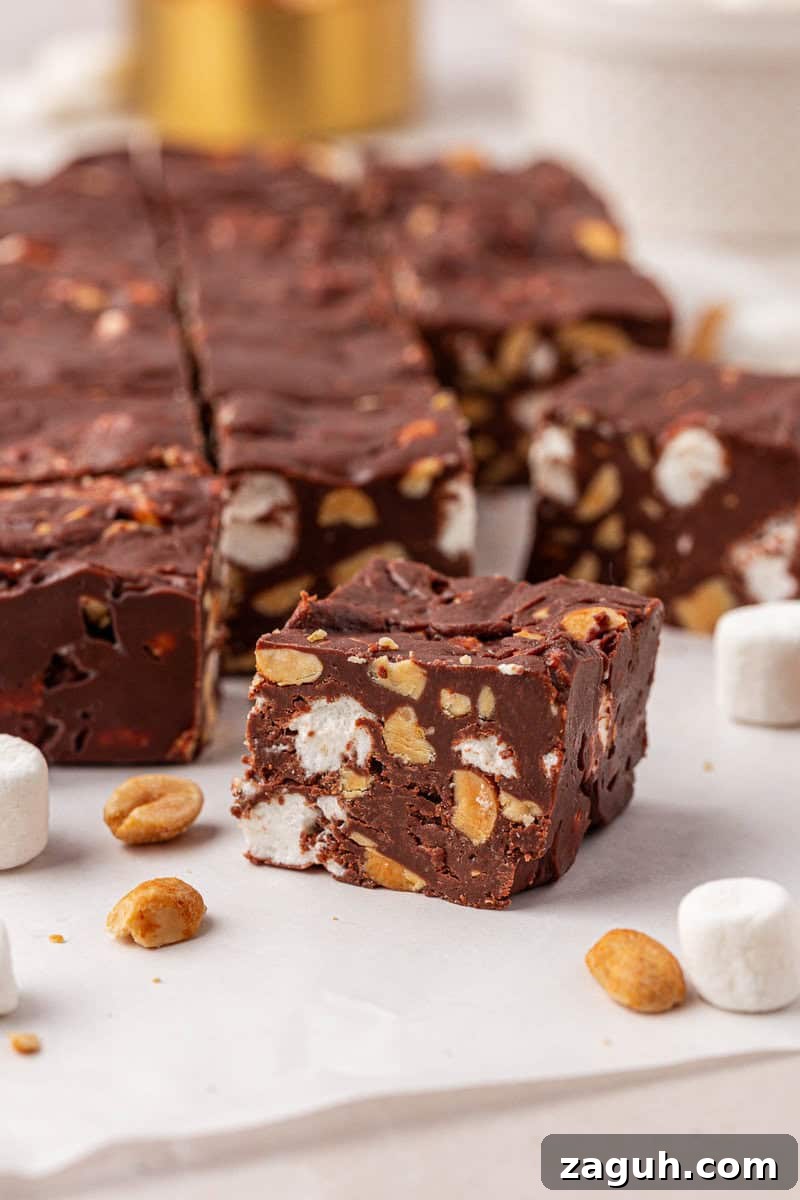 A close-up of chocolate fudge squares with visible peanuts and mini marshmallows inside, surrounded by a few loose peanuts and marshmallows on a white surface.