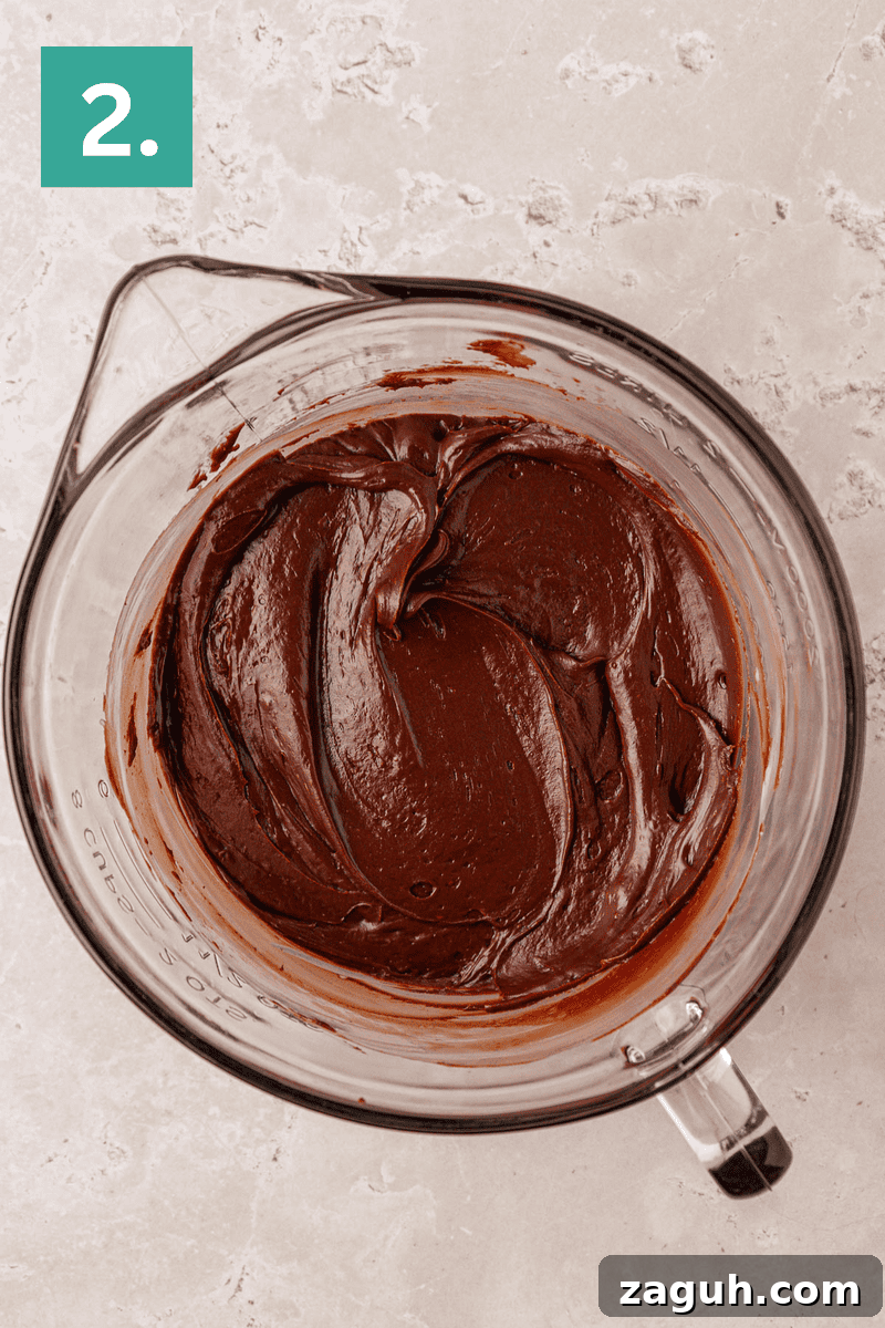 A glass mixing bowl filled with smooth, thick chocolate batter sits on a light-colored countertop. A green square with the number 2 is in the top left corner of the image.