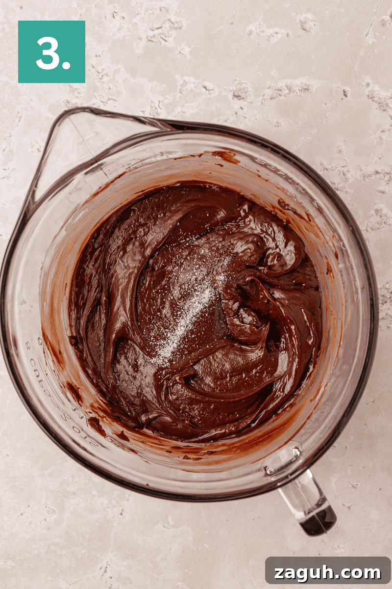 A glass mixing bowl filled with thick chocolate batter and a sprinkle of salt on top, placed on a light beige surface. A green box with the number 3 is in the upper left corner.