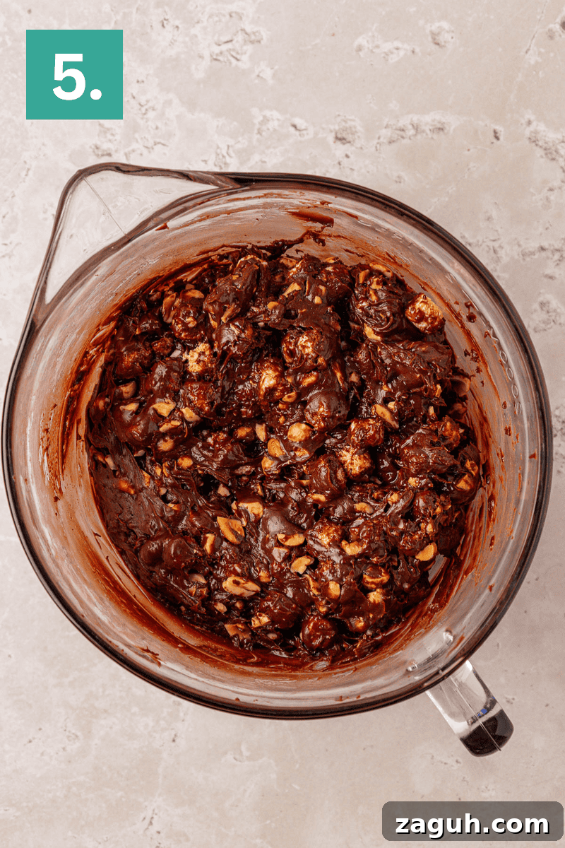 A glass mixing bowl filled with chocolate batter mixed with nuts and marshmallows sits on a light countertop. A green box with the number 5 is in the top left corner.