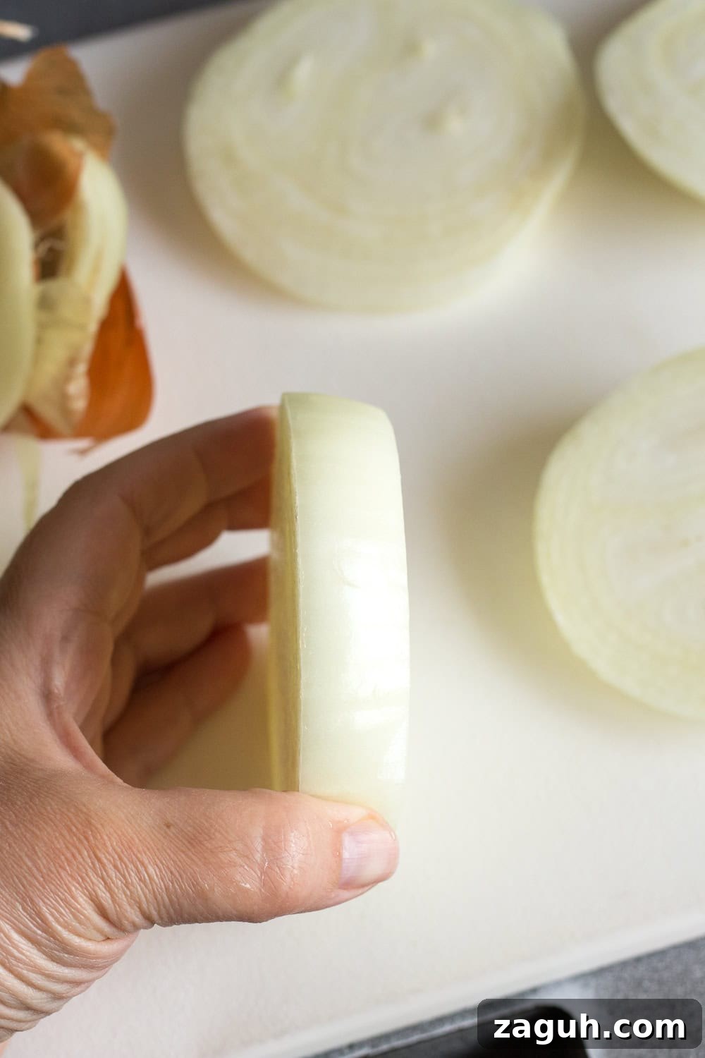 Grilled Onions Recipe: Whole30, Paleo & Gluten-Free Side Dish 4 A hand holding up a large slice of an onion on a white cutting board. Other slices of onion lay on the cutting board around it.