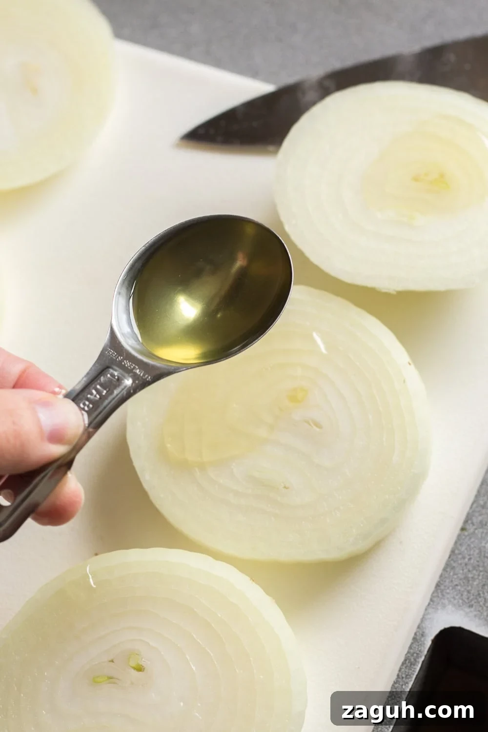 Grilled Onions Recipe: Whole30, Paleo & Gluten-Free Side Dish 5 A silver tablespoon pouring oil on a large slice of an onion on a white cutting board.