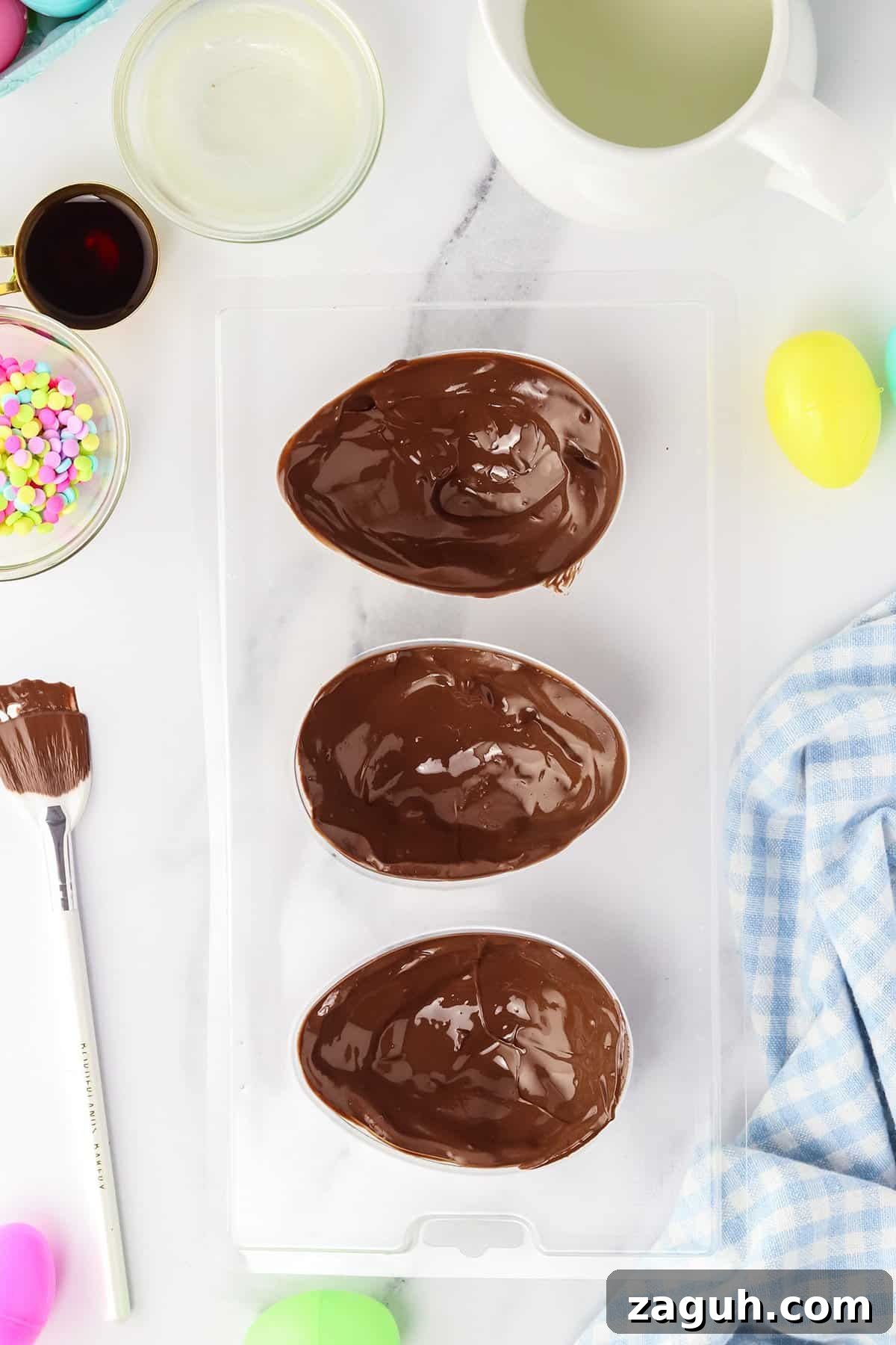 How to Make Copycat Cadbury Creme Egg Candy at Home 5 Three chocolate Easter egg halves in a mold, surrounded by ingredients and tools for making homemade Copycat Creme Egg candy.